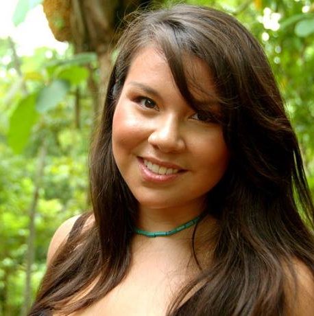 Ashley is a Mohawk woman in her 30s. She smiles brightly in front of a hedge. She wears a green choker style necklace and her brown hair falls across her forehead and across her shoulders.