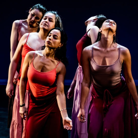 Five dancers are close together and softly press their neck and chest forward with closed eyes. They wear tank tops and skirts in varying shades of orange, red and burgundy.