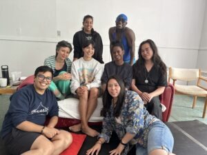8 people sit on, in front and stand behind a red couch in an art studio with white walls. Persons who are Indigenous, Black, Afrodescendant, Asian, cis gendered, non-binary and trans smile for a group photo.